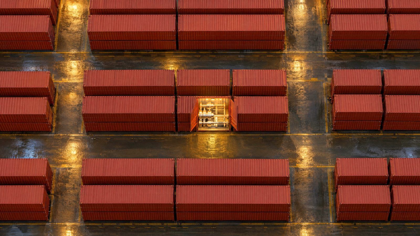 Rows of identical red shipping containers fill a wet port yard at night while a single open one shows precision machinery lit warm inside.